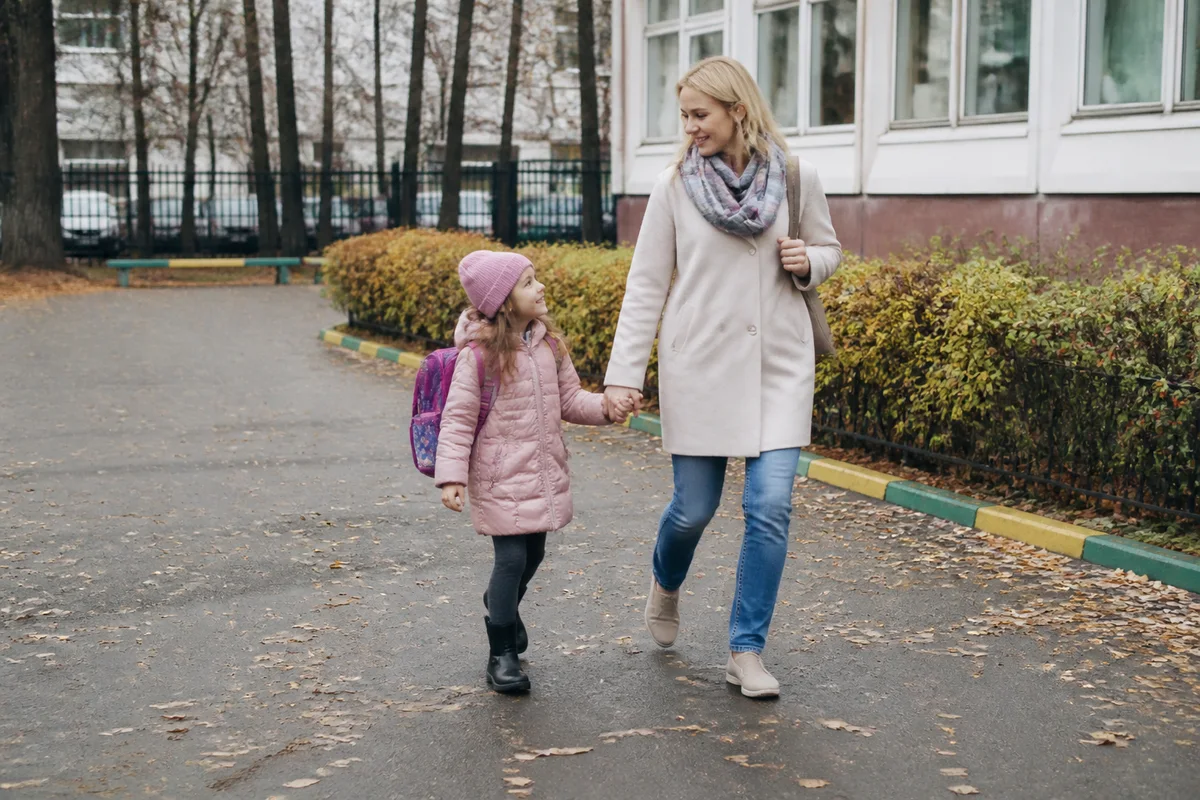 Шестилетняя девочка с новым рюкзаком идёт с мамой к школе поздней осенью
