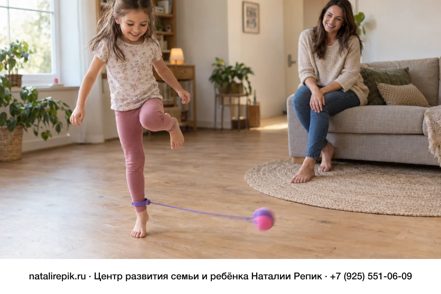 Девочка лет шести босиком занимается с нейроскакалкой в гостиной: шнур с розовым шариком закреплён на лодыжке, девочка улыбается и стоит на одной ноге; мама наблюдает с дивана
