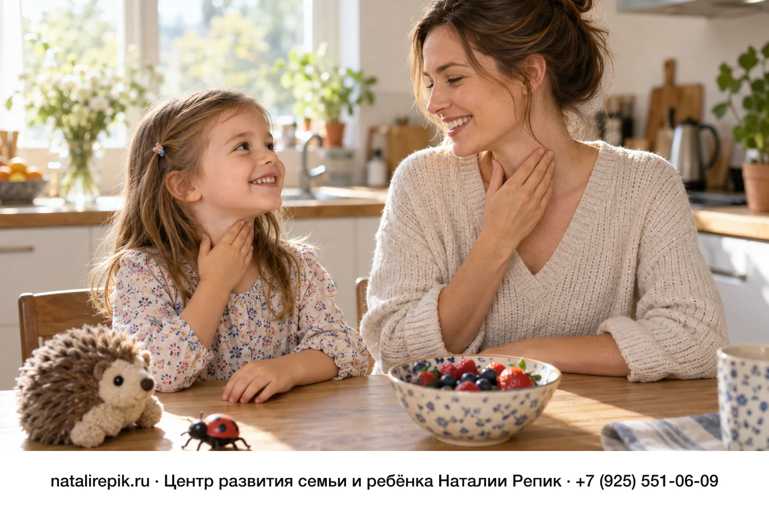 Мама и дочка лет пяти сидят рядом за кухонным столом, каждая приложила ладонь к своему горлу, чтобы почувствовать голос, обе улыбаются; на столе мягкий ёжик, игрушечная божья коровка и миска с ягодами
