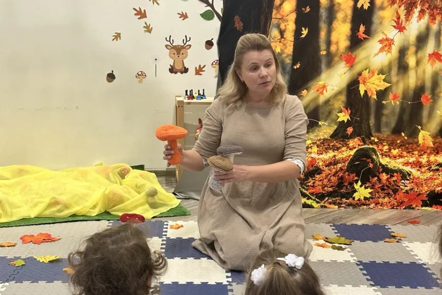 Дети сидят спиной к камере и слушают ведущую, которая показывает вязаные грибы