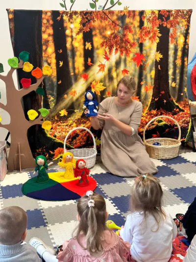 Ведущая показывает куклу в синем платье, рядом куклы-времена года на цветном круге и корзинки