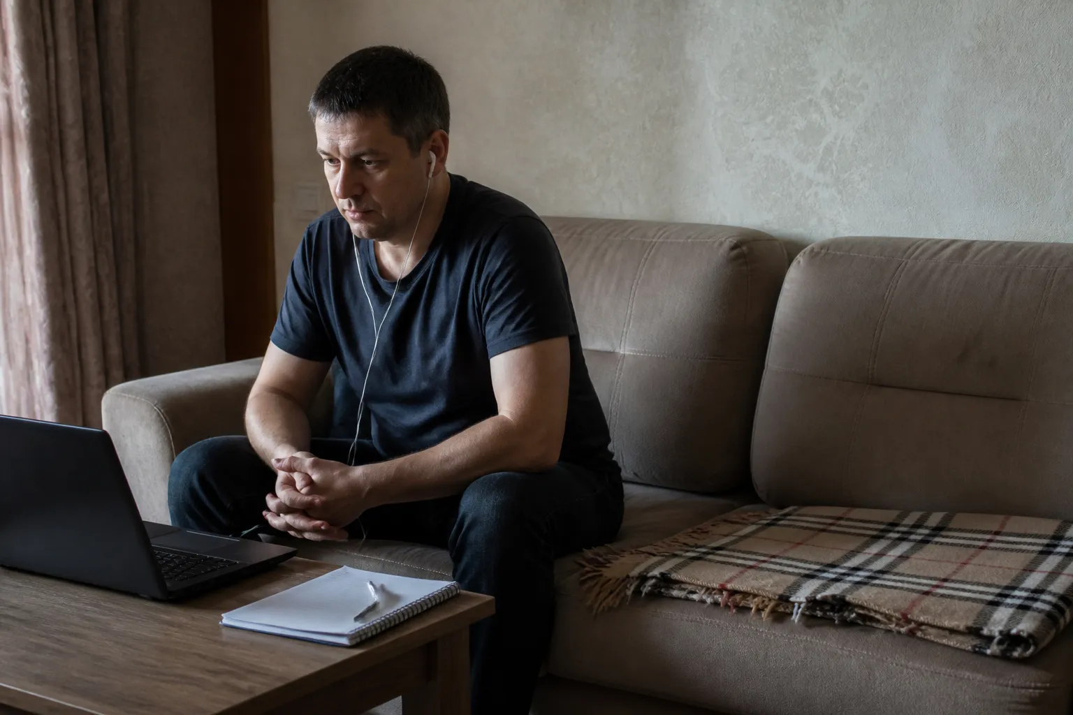 Мужчина дома на диване с ноутбуком и наушниками на сессии, вторая половина дивана пуста: прийти можно одному